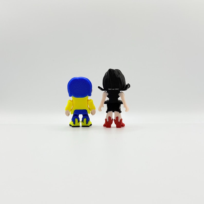 K&ouml;nnte beinhalten: Zwei kleine Figuren, eine mit blauen Haaren, einer gelben Jacke und blauen Hosen, und die andere mit schwarzen Haaren und einem schwarzen Kleid, stehen nebeneinander. Die Figuren stehen vor wei&szlig;em Hintergrund.