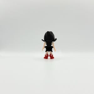 May include: A small figurine of a person viewed from the back. The figure has black hair, a black top, and a black skirt. The figure is wearing red boots. The figurine is standing on a white surface.