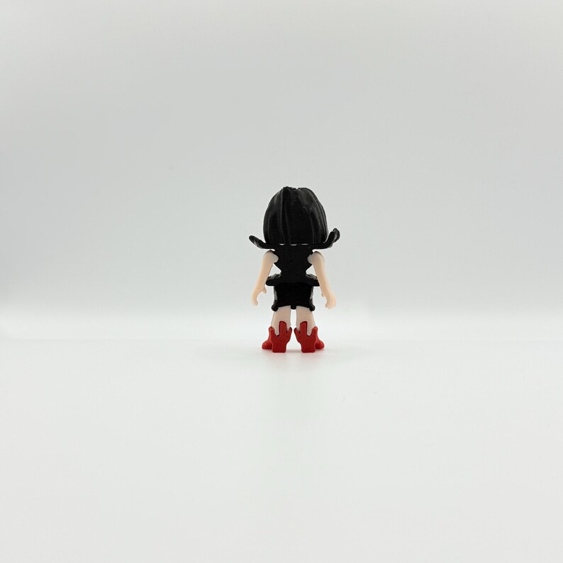 May include: A small figurine of a person viewed from the back. The figure has black hair, a black top, and a black skirt. The figure is wearing red boots. The figurine is standing on a white surface.