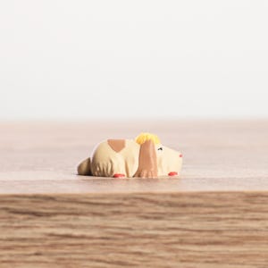 K&ouml;nnte beinhalten: Kleine, beige Hundefigur mit braunen und gelben Akzenten, liegend. Der Hund hat rote Details an den Pfoten und einen kleinen braunen Fleck auf dem R&uuml;cken. Die Figur liegt auf einer Holzoberfl&auml;che.