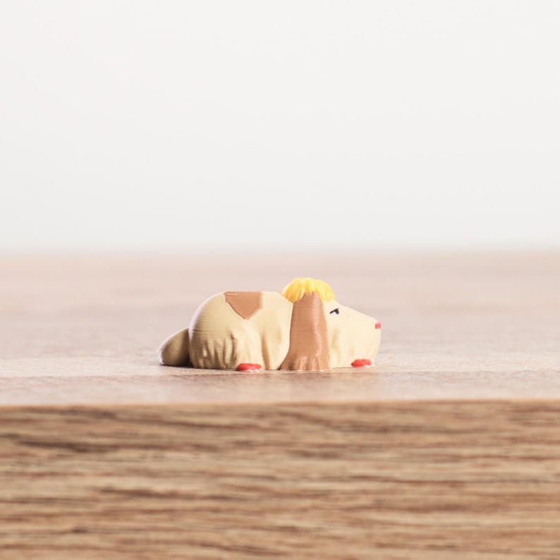 K&ouml;nnte beinhalten: Kleine, beige Hundefigur mit braunen und gelben Akzenten, liegend. Der Hund hat rote Details an den Pfoten und einen kleinen braunen Fleck auf dem R&uuml;cken. Die Figur liegt auf einer Holzoberfl&auml;che.