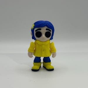 May include: A figurine of a character with blue hair, wearing a yellow coat, blue pants, and yellow boots. The character has large black eyes and a small flower in their hair. The figurine is standing against a white background.