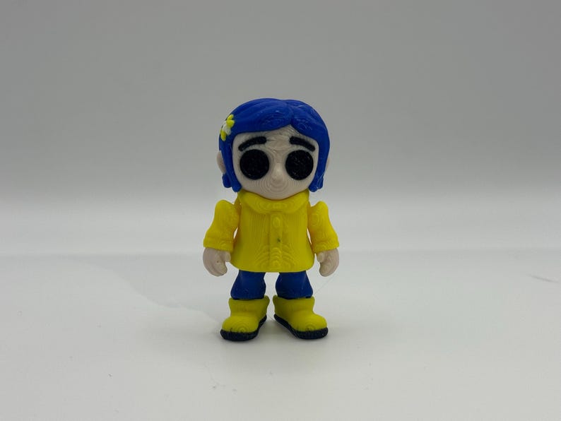 May include: A figurine of a character with blue hair, wearing a yellow coat, blue pants, and yellow boots. The character has large black eyes and a small flower in their hair. The figurine is standing against a white background.