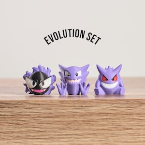 May include: A set of three purple and black figurines, representing a character evolution set. The figurines are displayed on a wooden surface, with the text "EVOLUTION SET" above them. The figurines have distinct facial features and poses.