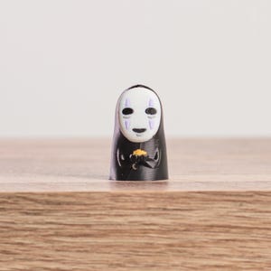 K&ouml;nnte beinhalten: Kleine, schwarze Figur der Figur Ohngesicht aus dem Zeichentrickfilm Spirited Away. Die Figur hat ein wei&szlig;es, maskenartiges Gesicht mit lila Akzenten und h&auml;lt ein kleines, goldenes Objekt. Auf einer Holzoberfl&auml;che stehend.