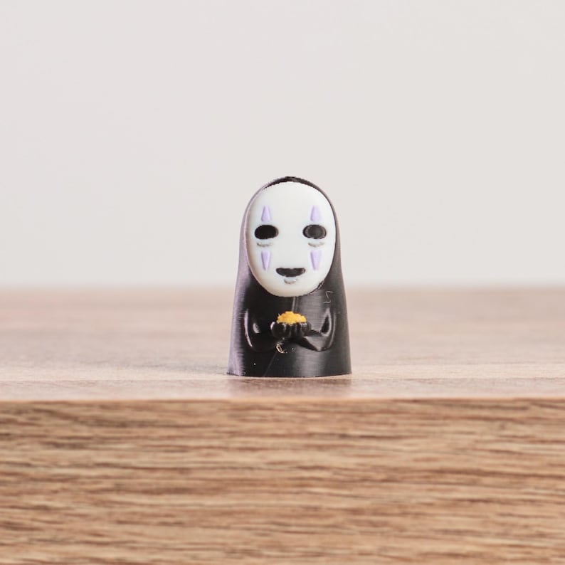 K&ouml;nnte beinhalten: Kleine, schwarze Figur der Figur Ohngesicht aus dem Zeichentrickfilm Spirited Away. Die Figur hat ein wei&szlig;es, maskenartiges Gesicht mit lila Akzenten und h&auml;lt ein kleines, goldenes Objekt. Auf einer Holzoberfl&auml;che stehend.