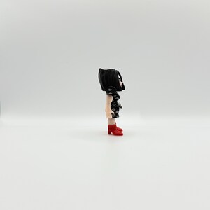 May include: A small figurine of a person with black hair and a black dress with white diamond shapes. The figurine is wearing red high-heeled shoes. The figure is standing on a white surface.
