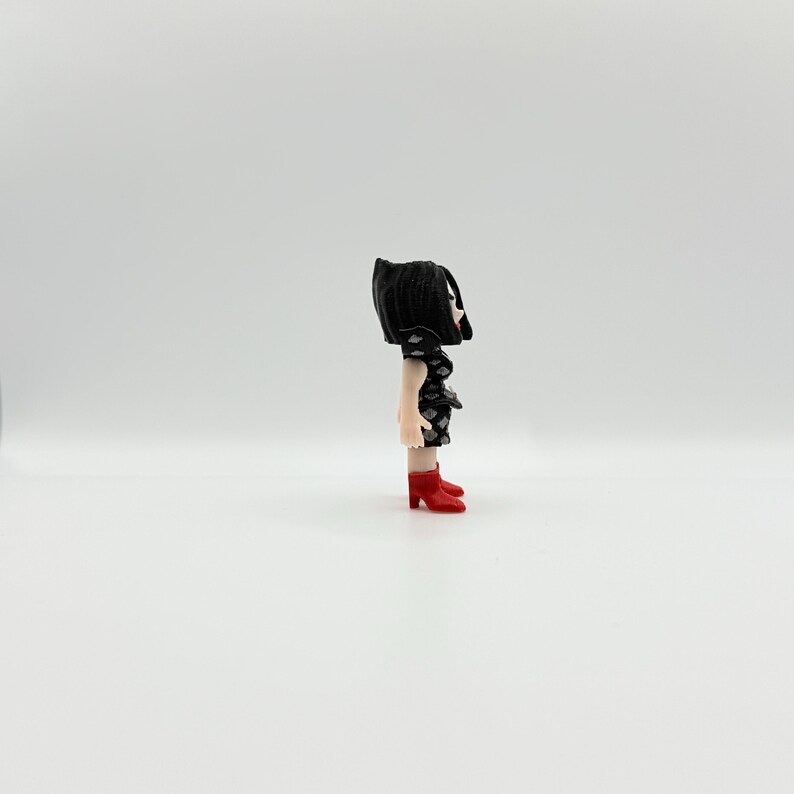 May include: A small figurine of a person with black hair and a black dress with white diamond shapes. The figurine is wearing red high-heeled shoes. The figure is standing on a white surface.