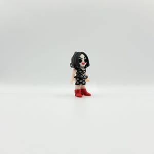 May include: A small figurine of a person with black hair and a black and white patterned dress. The figurine is wearing red boots. The figure has a stylized face with red lips and black eyes.