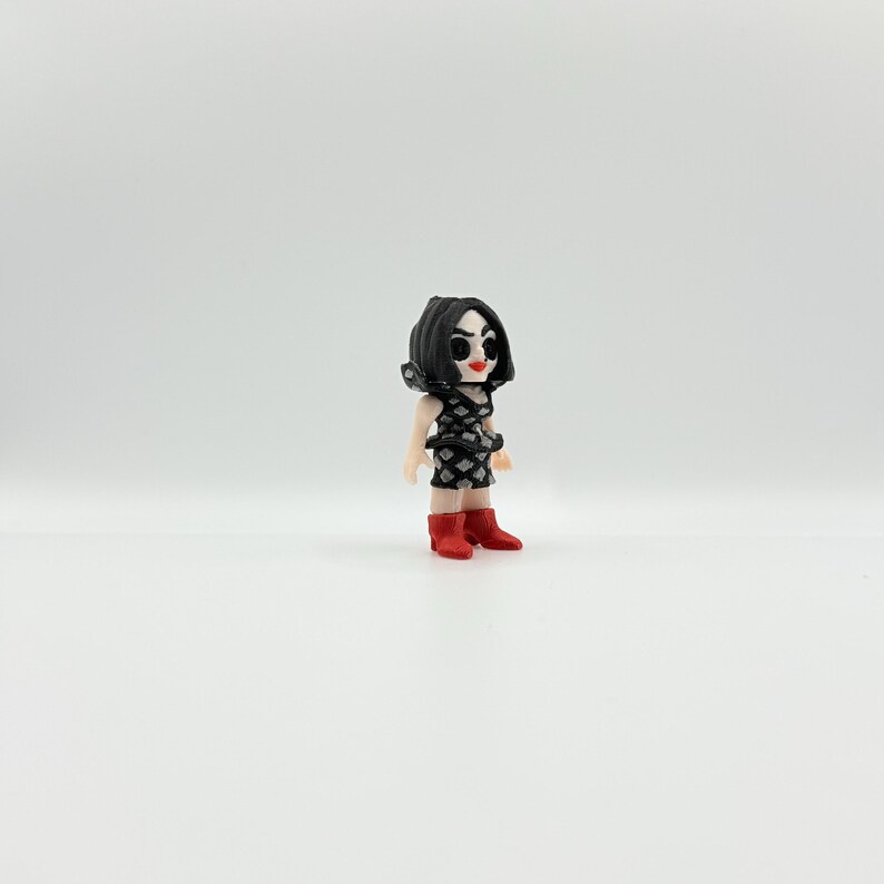 May include: A small figurine of a person with black hair and a black and white patterned dress. The figurine is wearing red boots. The figure has a stylized face with red lips and black eyes.