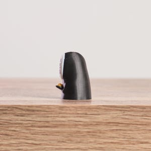 K&ouml;nnte beinhalten: Kleine, schwarze Figur mit wei&szlig;em Gesicht und einem kleinen, goldenen Gegenstand in der Hand. Die Figur steht auf einer Holzoberfl&auml;che, mit einem schlichten wei&szlig;en Hintergrund.