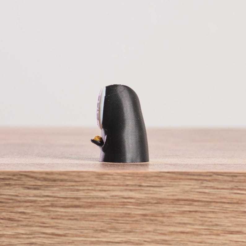K&ouml;nnte beinhalten: Kleine, schwarze Figur mit wei&szlig;em Gesicht und einem kleinen, goldenen Gegenstand in der Hand. Die Figur steht auf einer Holzoberfl&auml;che, mit einem schlichten wei&szlig;en Hintergrund.