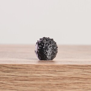May include: A small, black, spherical object with a textured, bumpy surface. The object is centered on a wooden surface, with a plain white background. The object appears to be a decorative element or a small sculpture.