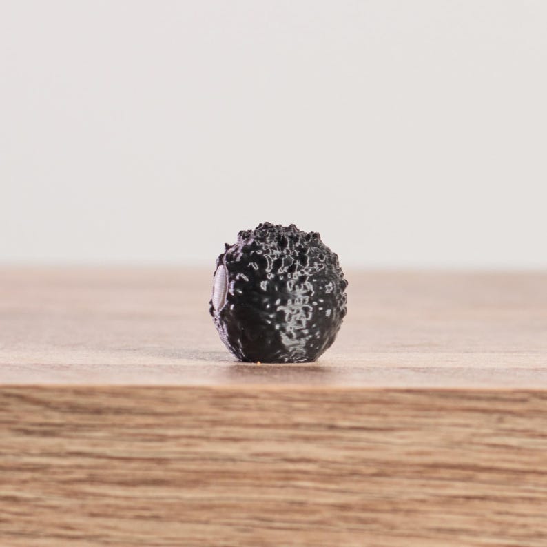 May include: A small, black, spherical object with a textured, bumpy surface. The object is centered on a wooden surface, with a plain white background. The object appears to be a decorative element or a small sculpture.