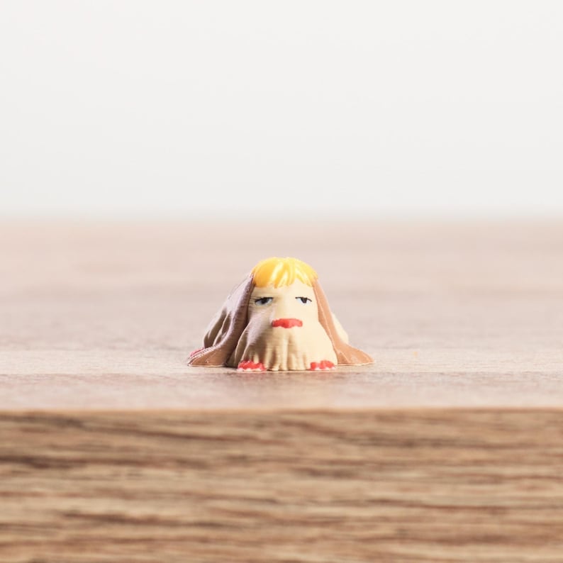 K&ouml;nnte beinhalten: Eine kleine, skurrile Figur eines Charakters mit gelben Haaren, einem beigen Gesicht und einem roten Mund. Die Figur ist in einem braunen Tuch drapiert, mit roten Akzenten am unteren Rand. Die Figur steht auf einer Holzoberfl&auml;che.