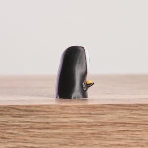 K&ouml;nnte beinhalten: Kleine, schwarz-wei&szlig;e Pinguin-Figur mit gelbem Schnabel und F&uuml;&szlig;en. Der Pinguin steht auf einer Holzoberfl&auml;che. Die Figur ist ein dekoratives Objekt.
