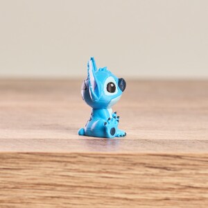 Peut inclure: Petite figurine bleue clair du personnage Disney Stitch, avec de grands yeux noirs et des oreilles pointues. La figurine est pos&eacute;e sur une surface en bois.