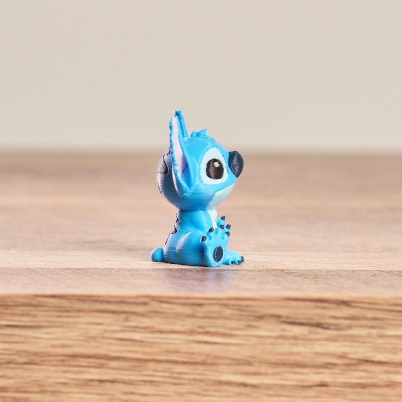 Peut inclure: Petite figurine bleue clair du personnage Disney Stitch, avec de grands yeux noirs et des oreilles pointues. La figurine est pos&eacute;e sur une surface en bois.
