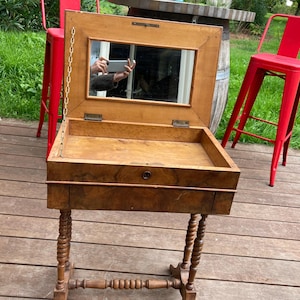 Peut inclure: Une coiffeuse vintage en bois avec un dessus à charnière et un petit miroir. La table a un tiroir et est soutenue par des pieds tournés ornés. Le dessus est ouvert, révélant un compartiment de rangement. Une chaîne maintient le dessus ouvert.