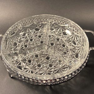 Peut inclure: Plat de service en verre transparent à trois compartiments, avec un motif en forme d'éclat d'étoile. Le plat repose dans un support en filigrane en métal argenté avec deux poignées. Une pièce décorative pour servir des collations ou des apéritifs.