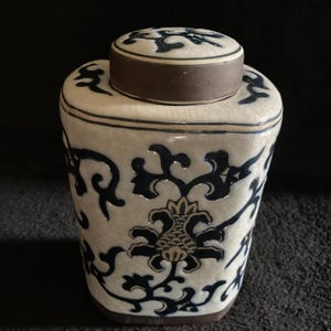 Peut inclure: Un pot en céramique blanc et bleu avec un couvercle. Le pot présente un motif floral et de volutes bleu sur fond blanc. Le couvercle est marron avec un motif floral bleu. Le pot a une forme rectangulaire aux bords arrondis.