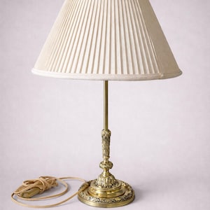 Peut inclure: Lampe de table de style vintage avec un abat-jour plissé crème. La lampe a un pied et une tige en laiton avec des détails ornés. Le cordon est de couleur beige clair.