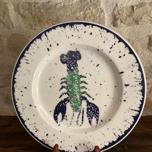 Peut inclure: Assiette en céramique blanche avec un bord bleu marine et un motif éclaboussé. L'assiette présente une illustration de homard vert et bleu marine au centre. L'assiette est présentée sur un présentoir en bois contre un mur en pierre.