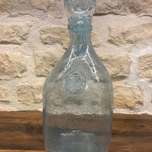 Puede incluir: Una botella de vidrio transparente de color azul claro con un tapón redondo. La botella tiene un cuerpo bulboso y un cuello largo. El vidrio tiene una apariencia texturizada y con burbujas. Un emblema circular está grabado en la parte delantera. La botella está sobre una superficie de madera.
