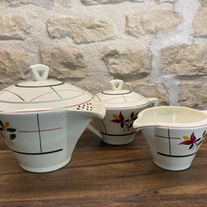 Peut inclure: Un service à thé vintage de couleur crème comprenant une théière, un sucrier et un pot à crème. L'ensemble présente un motif géométrique avec des accents floraux rouges, verts et jaunes. La théière a un couvercle et un bec verseur.