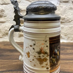 Peut inclure: Une chope à bière vintage avec un couvercle et une poignée en étain à charnière. Le corps en céramique blanche présente des motifs floraux dorés et une scène peinte. Des bandes marron entourent la base et le haut. La chope mesure environ 20 cm de haut.