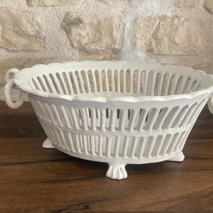 Può includere: Un cestino ovale in ceramica bianca con un design traforato. Il cestino ha due manici ad anello e poggia su tre piedini. Il cestino è esposto su una superficie di legno scuro.