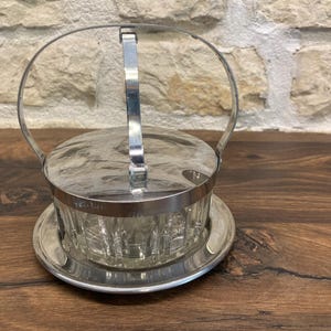 Peut inclure: Un sucrier vintage argenté avec un insert en verre, un couvercle et une poignée. Le bol repose sur une soucoupe argentée assortie. Le design présente un corps en verre clair et texturé et une finition métallique brillante.
