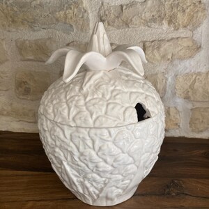 Puede incluir: Un recipiente blanco de cerámica con forma de piña y tapa. El recipiente tiene una superficie texturizada y un asa con forma de flor en la parte superior. El recipiente está sobre una superficie de madera.