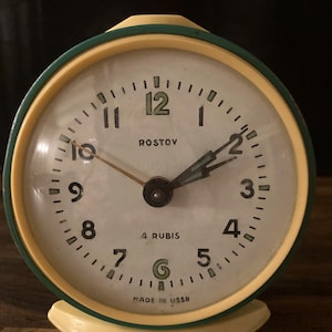 Peut inclure: Un réveil vintage rond avec un cadran crème et un cadre crème et vert. Le cadran de l'horloge comporte des chiffres et des aiguilles noirs, avec le mot "ROSTOV" au-dessus du centre. Le texte "4 RUBIS" et "MADE IN USSR" est également visible.