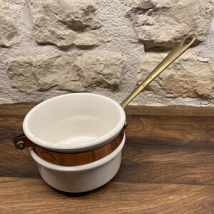 Peut inclure: Une petite casserole en émail blanc avec une bande de cuivre et une longue poignée en laiton. La casserole est posée sur une surface en bois et un fond de mur en pierre. La poignée mesure environ 20 cm.
