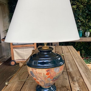Op de afbeelding: Een decoratieve tafellamp met een crèmekleurige lampenkap. De voet is donkerblauwgroen met een bloemenontwerp in oranje, rood en blauw. De lamp staat op een houten tafel.