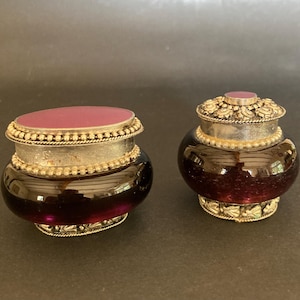 Pode incluir: Dois frascos de vidro antigos com detalhes em metal prateado. Os frascos têm um tom roxo profundo e são cobertos com tampas a condizer. As tampas são adornadas com uma pedra roxa central e trabalhos decorativos em metal. Os frascos são provavelmente para guardar pequenos objetos.