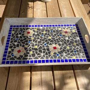 Puede incluir: Bandeja rectangular con diseño de mosaico. La bandeja tiene un marco de madera gris claro con asas. El mosaico presenta azulejos negros, grises, amarillos, rojos y azules dispuestos en un patrón floral.