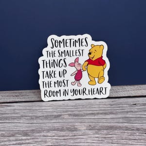 May include: A white sticker featuring Winnie the Pooh and Piglet holding hands. The yellow Pooh wears a red shirt, and Piglet is pink with a striped shirt. The sticker has the text: "Sometimes the smallest things take up the most room in your heart."