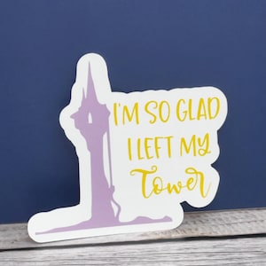 May include: A white sticker with a purple tower silhouette and yellow text that reads "I'M SO GLAD I LEFT MY Tower". The sticker is on a wooden surface with a dark blue background.