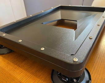 May include: A black rectangular platform with a recessed centre and rounded corners. The platform is mounted on four black suction cup feet. Silver screws secure the frame. The platform sits on a wooden surface.