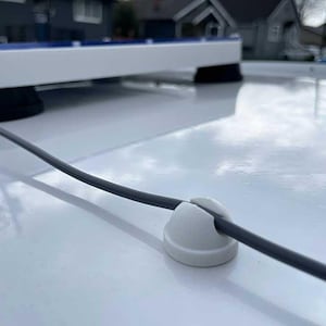 Op de afbeelding: Een witte kabelklem die een grijze kabel vastzet. De klem is bevestigd op een wit oppervlak, met een groter wit en blauw object op de achtergrond. De foto is buiten genomen.