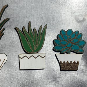 May include: Six wooden plant pot ornaments with different plant designs. The plants are painted in shades of green and blue. The pots are painted white with decorative details.