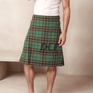 Puede incluir: Un kilt de cuadros verdes, marrones y amarillos con una correa de cuero negra y una hebilla. El kilt lo lleva puesto una persona de pie sobre una superficie de color marrón claro.