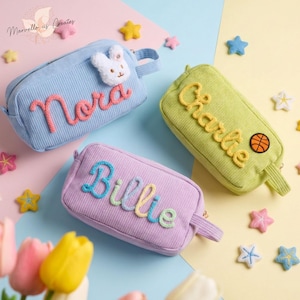 May include: Three rectangular corduroy pencil cases in light blue, lime green, and lavender. The blue case features a white plush bunny and the name Nora in pink. The green case has the name Charlie and a basketball patch. The lavender case has the name Billie in multi-coloured yarn.