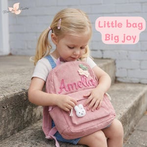 Peut inclure: Un sac à dos en velours côtelé rose avec le nom "Amelia" brodé. Il est orné d'un charm en forme de lapin blanc et de fleurs. Le texte "Little bag Big joy" est également visible.
