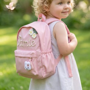 Peut inclure: Un sac à dos rose clair portant le prénom Jamie brodé en lettres cursives blanches, orné de deux fleurs crochetées blanches sur le dessus et d'une petite breloque lapin en peluche blanche attachée à la fermeture éclair de la poche avant.
