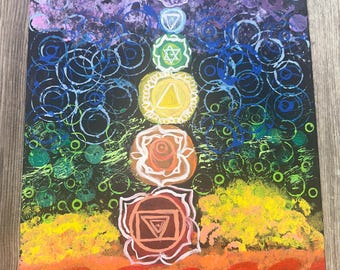 May include: A black canvas painting featuring a colourful depiction of the seven chakras. The chakras are represented by circles with symbols inside, and are arranged vertically in a column. The background is a dark purple, blue, green, yellow, and orange.