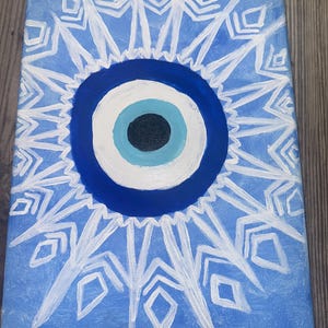 May include: A blue and white canvas painting with a white mandala design surrounding a blue evil eye. The evil eye has a white iris and a black pupil.