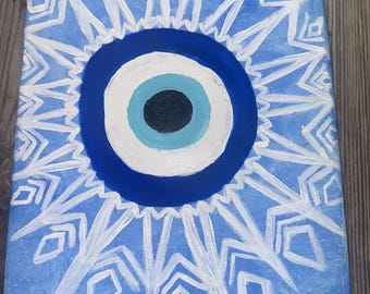 May include: A blue and white canvas painting featuring a white mandala design surrounding a blue evil eye. The evil eye has a white iris and a black pupil.